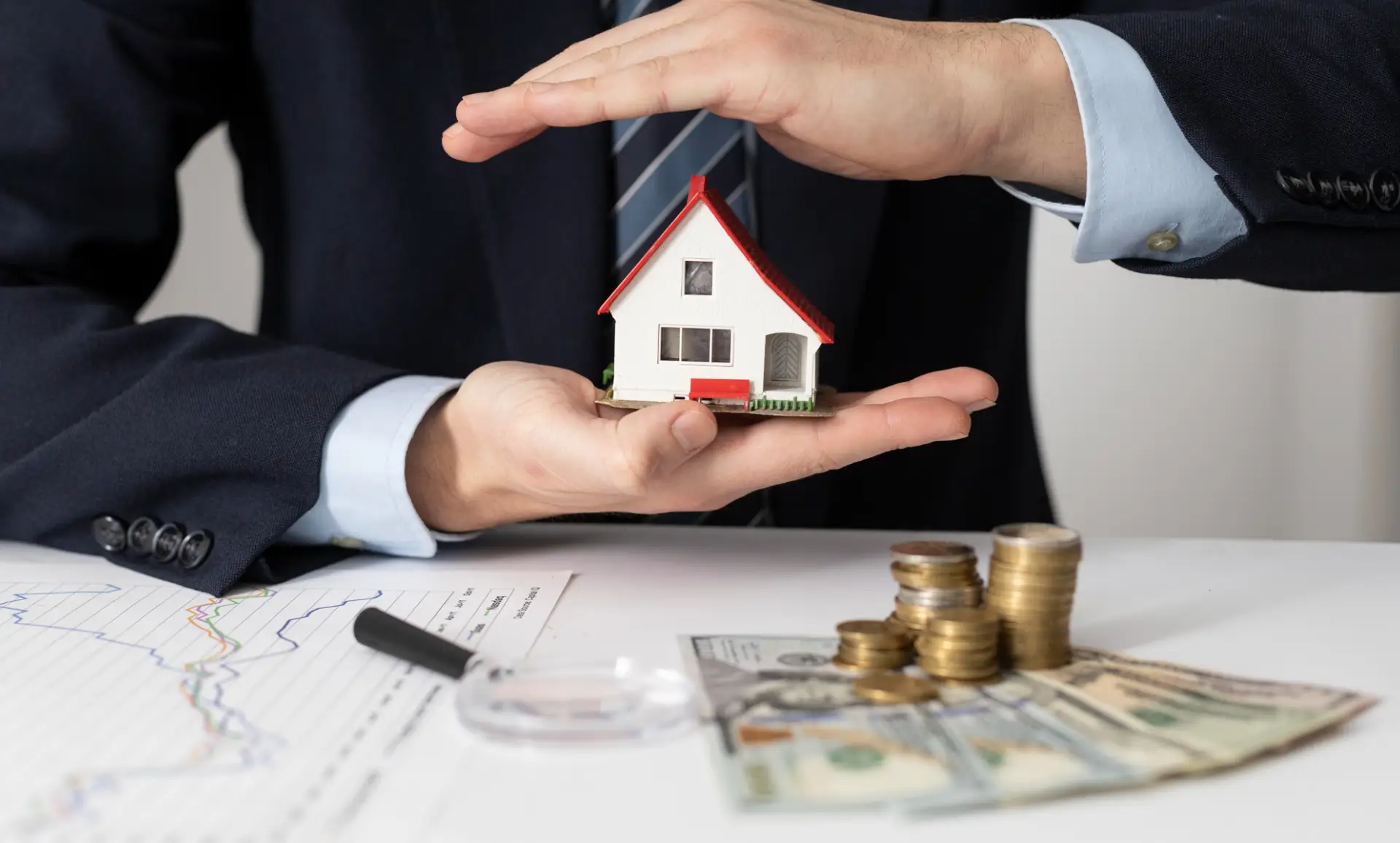 A businessman in a suit holds a small house model, symbolizing real estate investment, with money and charts in the background