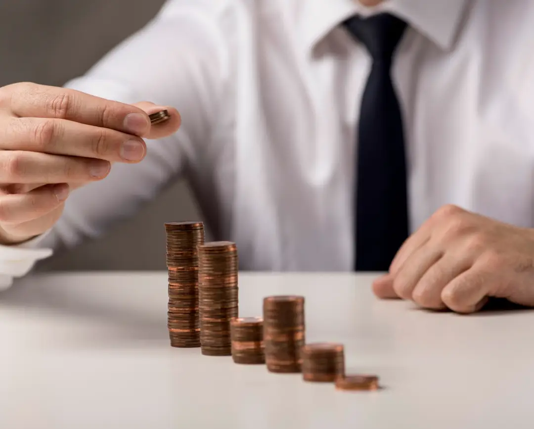 A man in a white shirt and tie stacks coins, illustrating financial growth, with several ascending towers of pennies in front of him