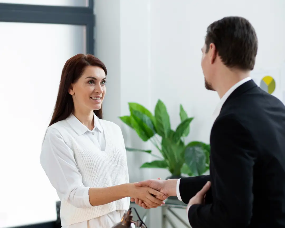 A professional setting with two individuals shaking hands, a desk with a coffee cup and papers, and greenery in the background