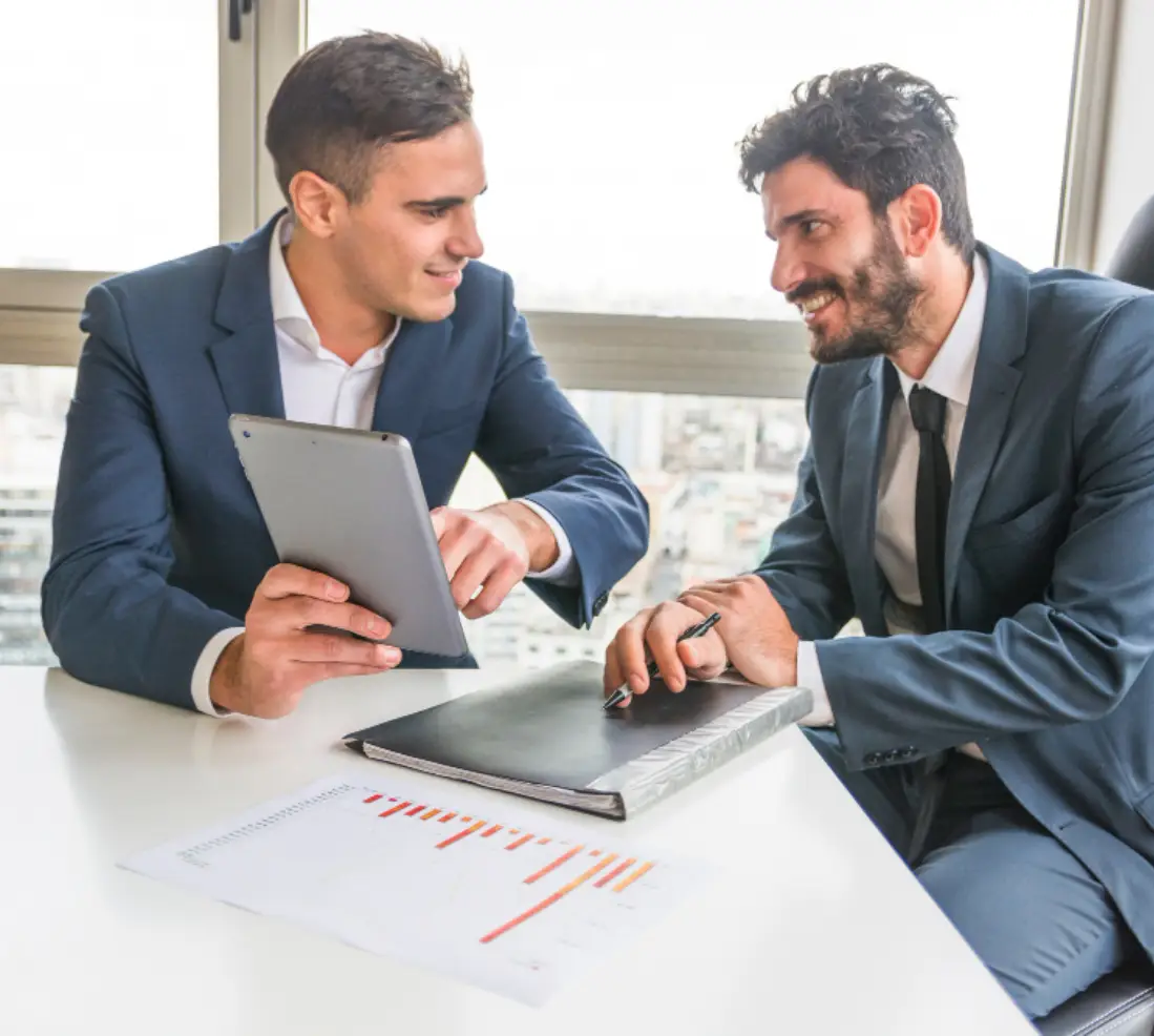 Two business professionals in formal attire are engaged in a discussion at a modern office table with a city skyline view, one using a tablet